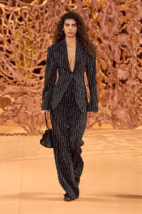 Model walking a runway in a long black pinstripe suit with a deep V, carrying a small black handbag.