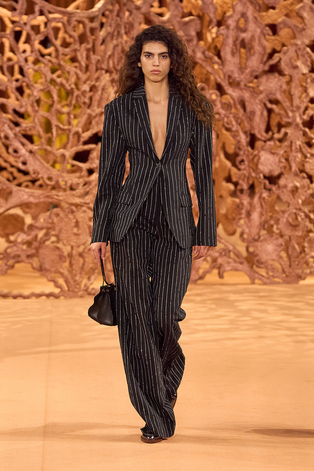 Model walking a runway in a long black pinstripe suit with a deep V, carrying a small black handbag.