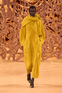 Model walks a runway wearing a mustard yellow oversized coat with a large scarf detail, paired with black-heeled patterned boots, against a sculptural brown backdrop.