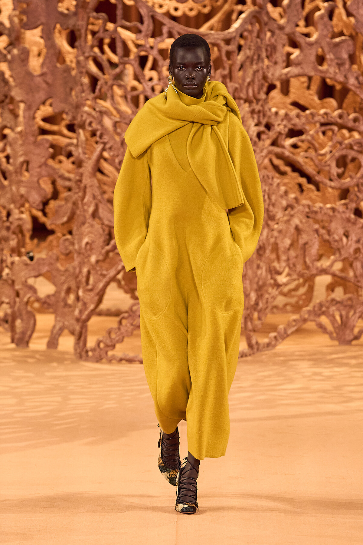 Model walks a runway wearing a mustard yellow oversized coat with a large scarf detail, paired with black-heeled patterned boots, against a sculptural brown backdrop.