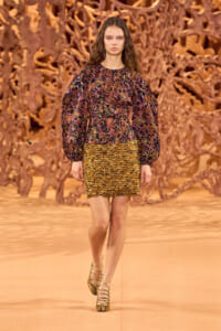 Model walking the runway in a long-sleeve, multicolor scale-pattern dress with a gold scalloped hem.