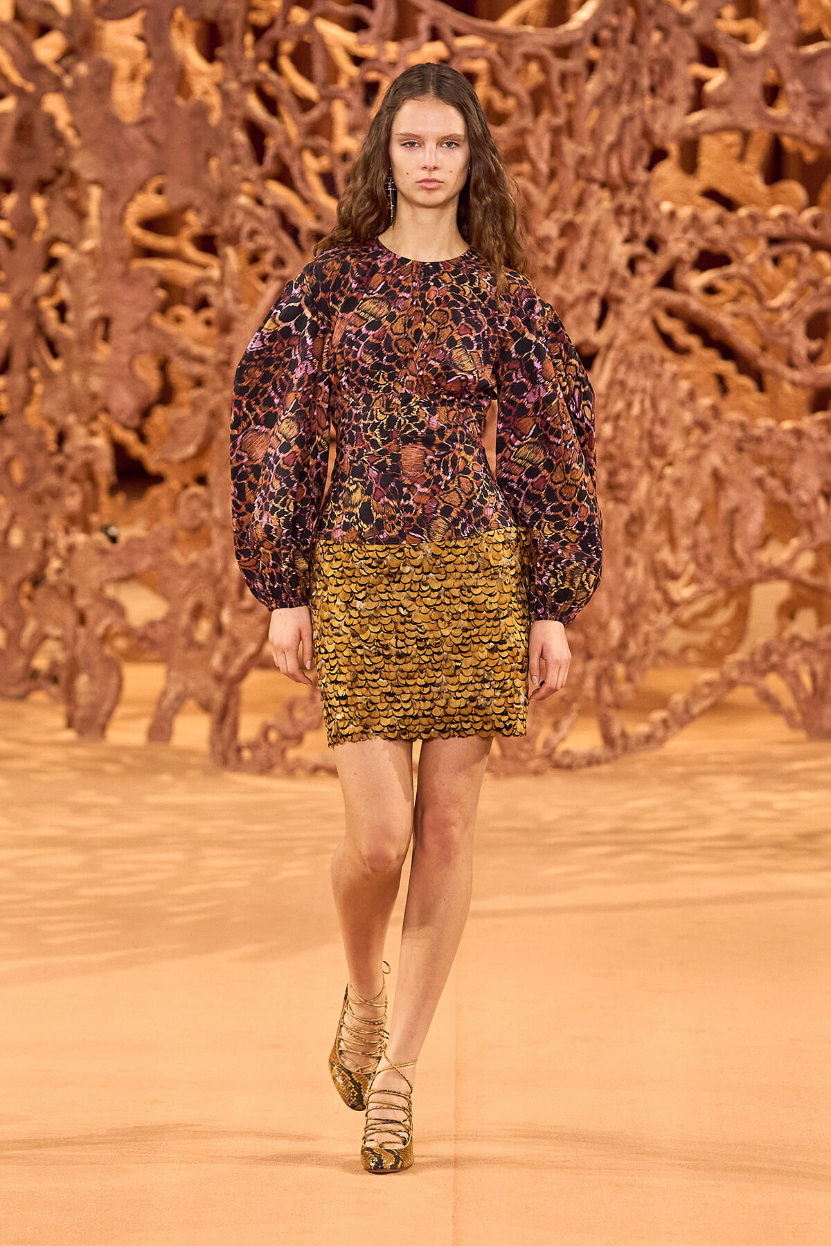 Model walking the runway in a long-sleeve, multicolor scale-pattern dress with a gold scalloped hem.