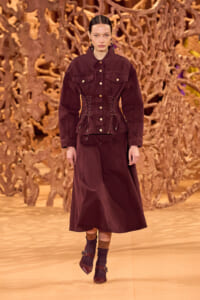 Model on a fashion runway wearing a burgundy/cranberry jacket with corset-style lacing and a matching long skirt, against a sculptural wooden backdrop.