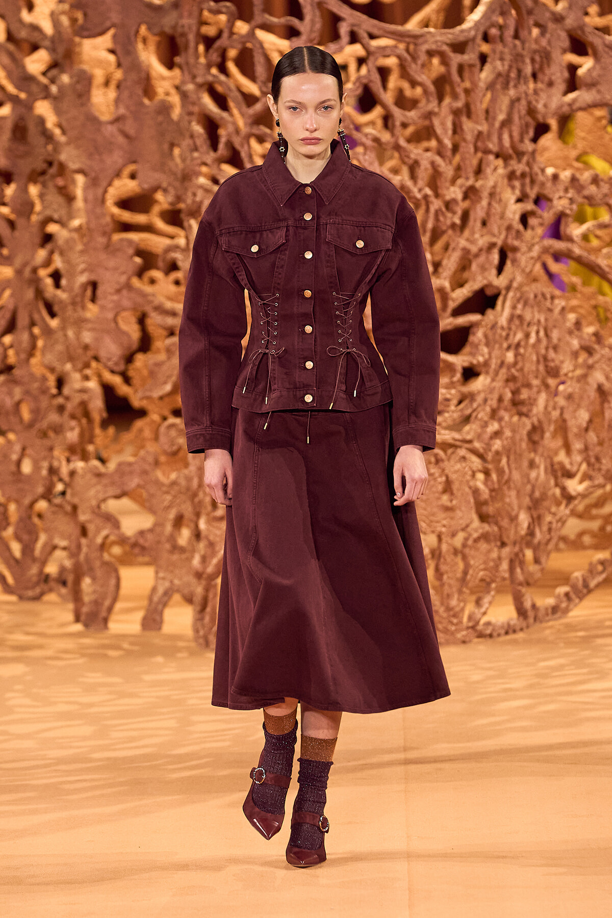 Model on a fashion runway wearing a burgundy/cranberry jacket with corset-style lacing and a matching long skirt, against a sculptural wooden backdrop.