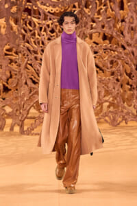 Model on a runway wearing a purple turtleneck, camel overcoat, and brown leather pants with an ornate backdrop.