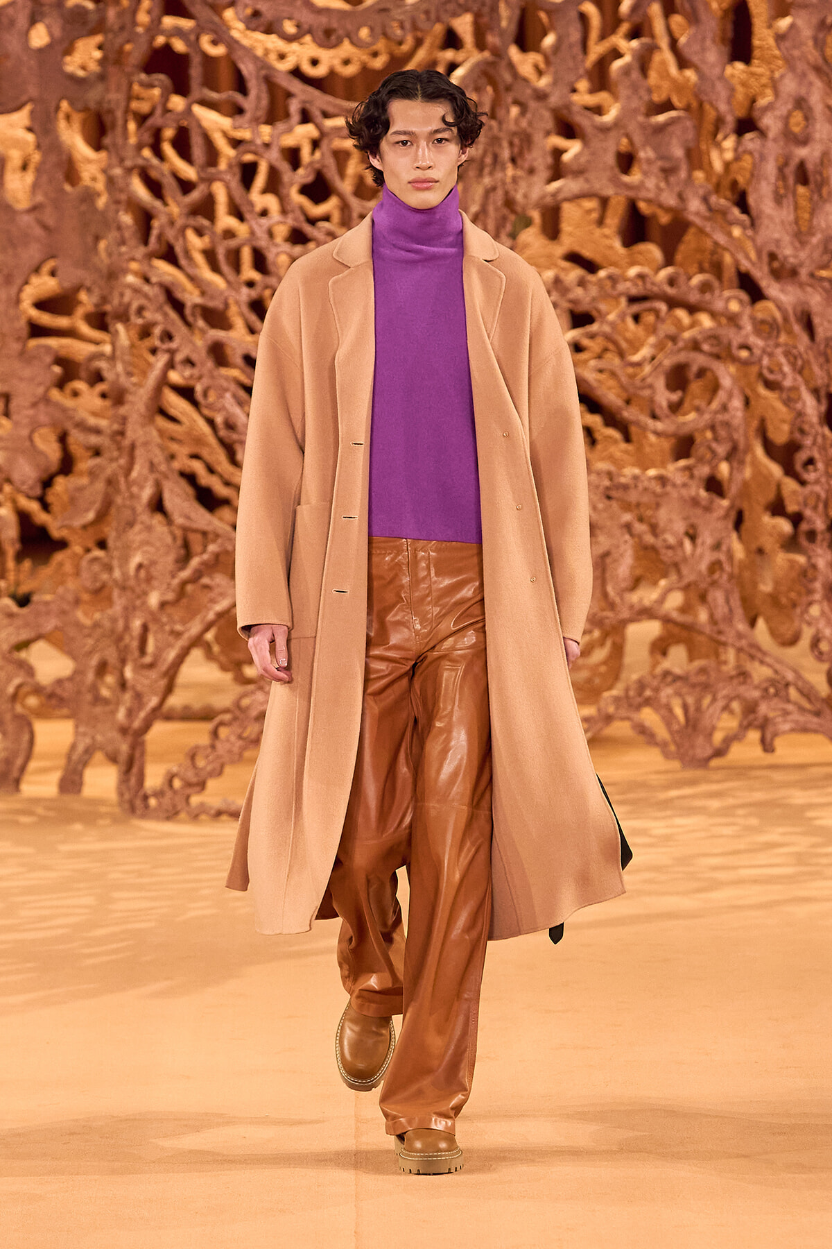 Model on a runway wearing a purple turtleneck, camel overcoat, and brown leather pants with an ornate backdrop.