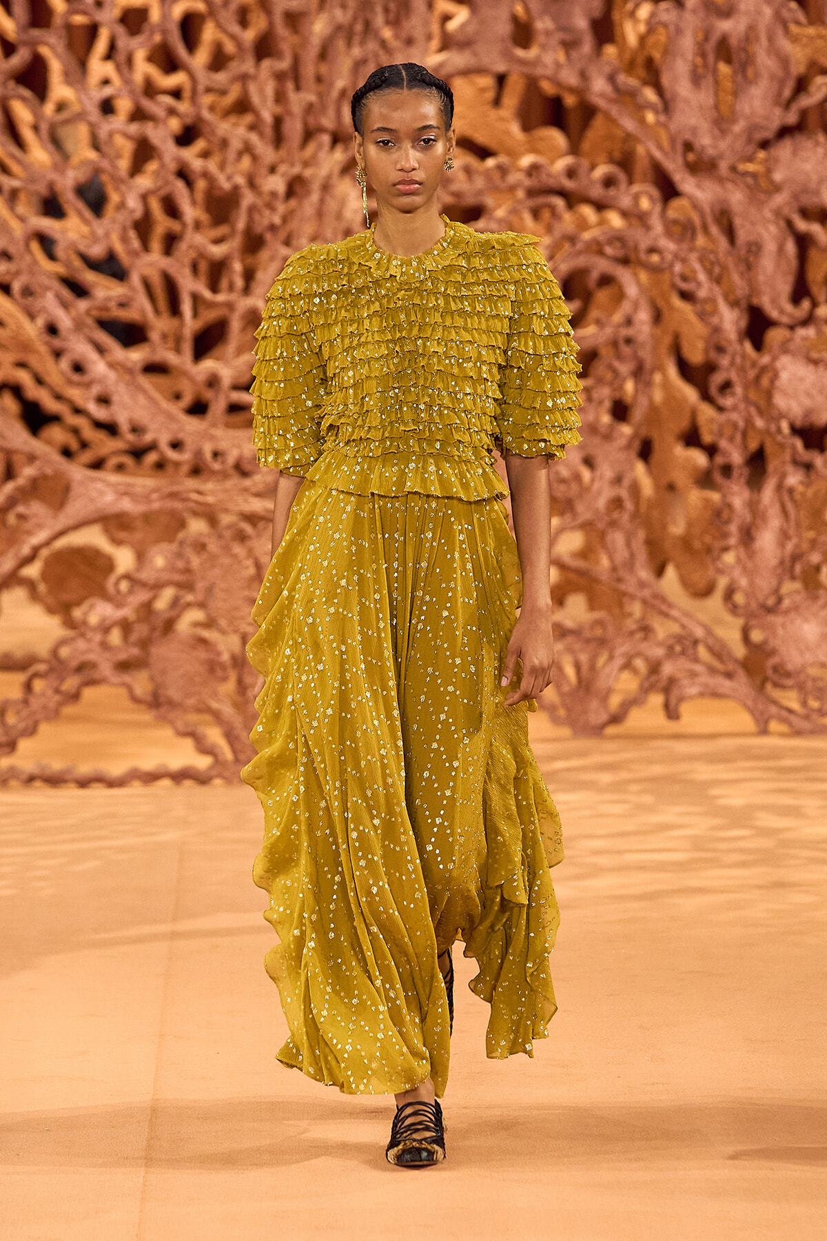 Model walking runway in mustard yellow ruffled dress with lace-like dots, black strappy heels; artistic orange backdrop.