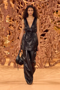 Model walks the runway in a sleeveless black leather V-neck top with matching wide-leg leather pants, carrying a black handbag.