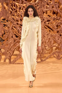 Model walking a runway in a cream ruffled gown with gold heels, against an intricate wooden backdrop.