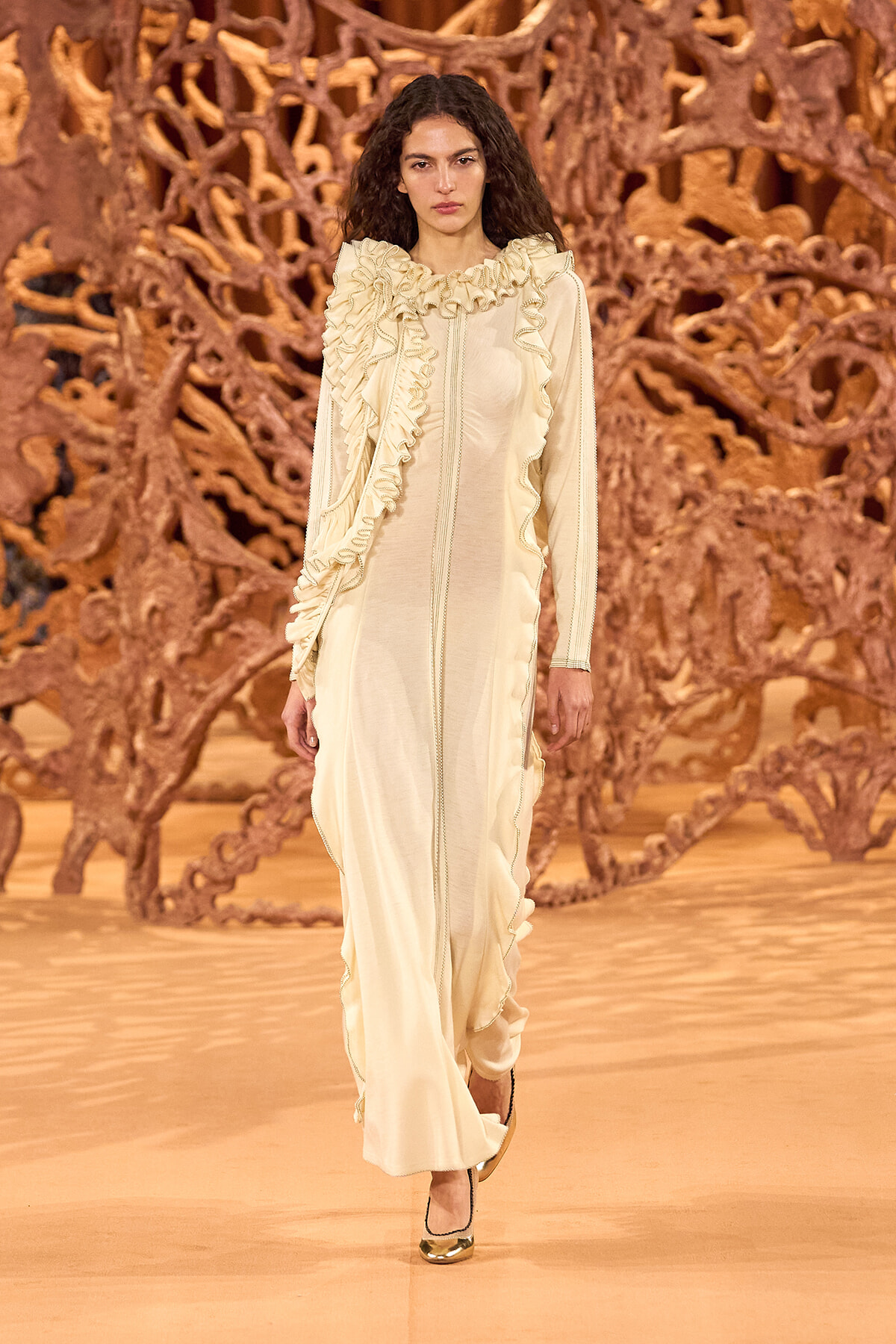 Model walking a runway in a cream ruffled gown with gold heels, against an intricate wooden backdrop.