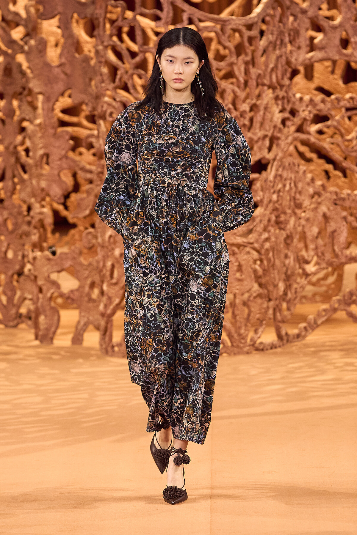 Model walking the runway in a long-sleeve, animal-print jumpsuit with puffed sleeves, belted at the waist, against a brown sculptural backdrop.