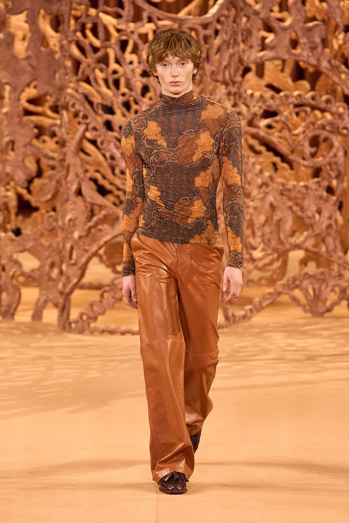Male model on a fashion runway wearing a rust-toned patterned turtleneck and brown leather trousers with a sculptural backdrop behind him.