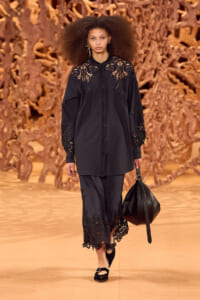 Model walks the runway in a black lace-detailed shirt dress, carrying a black handbag, against a warm, sculptural backdrop.