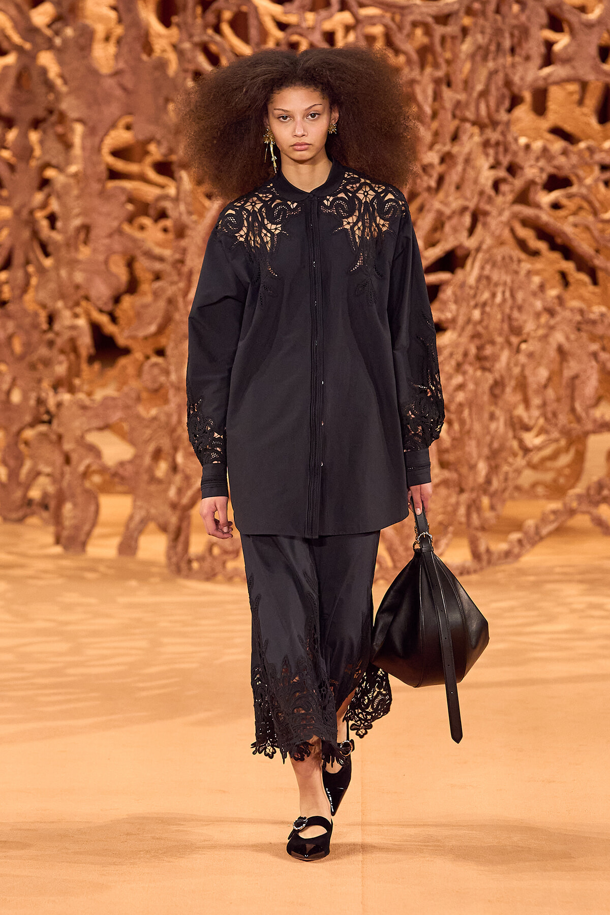 Model walks the runway in a black lace-detailed shirt dress, carrying a black handbag, against a warm, sculptural backdrop.