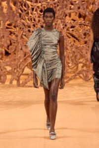Model walks the runway in a metallic silver one-shoulder ruched mini dress against an orange sculptural backdrop.