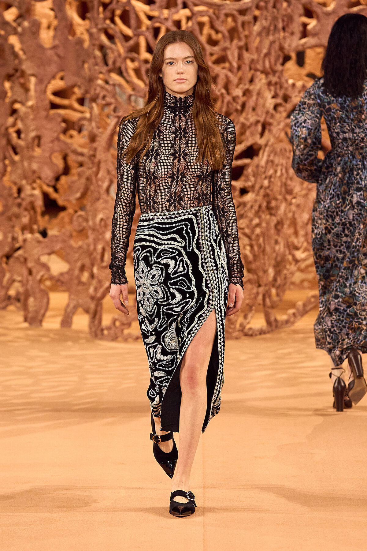 Model walking the runway in a black lace turtleneck top and a black-and-white patterned skirt with a high slit, against an orange sculptural backdrop.