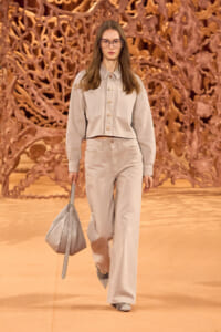 Model walks a runway wearing a beige cropped jacket and matching wide-leg pants, carrying a light gray handbag; copper-toned sculptural backdrop behind her.
