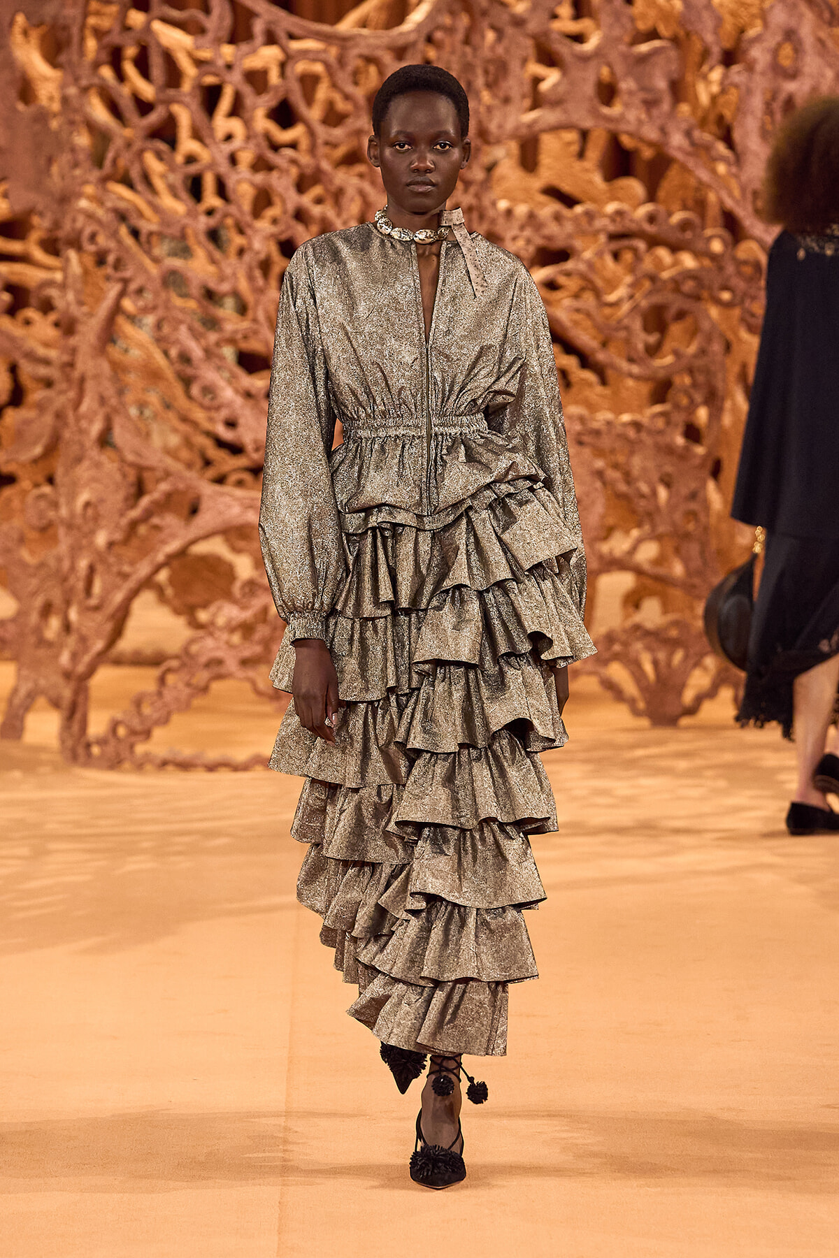 Model walking a metallic silver ruffled dress on a fashion runway with a sculptural wooden backdrop.