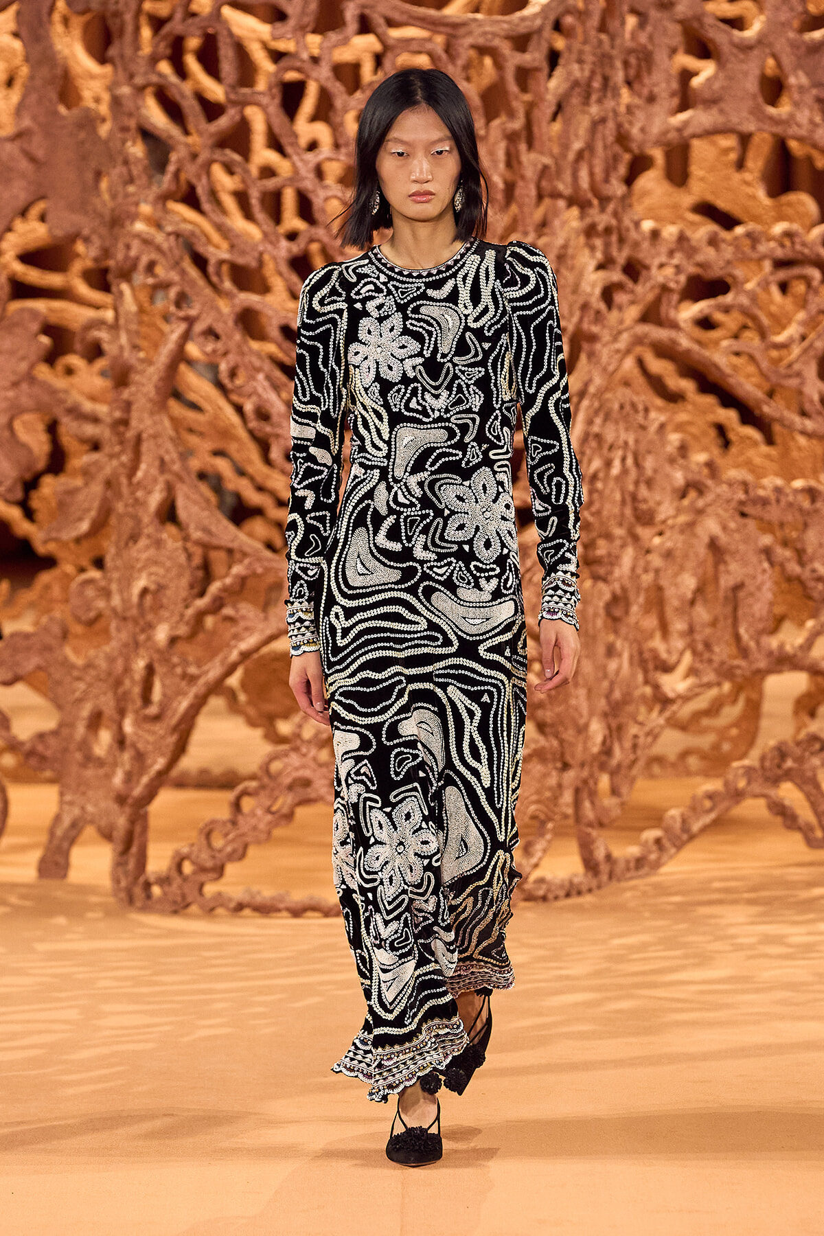 Model walks the runway in a long black-and-white patterned gown with swirling bead-like motifs, posed before a brown sculptural backdrop.