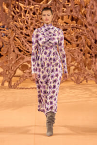 Model walking a runway in a long-sleeve purple floral jumpsuit with a high neck and gathered waist, paired with gray suede boots, orange sculptural backdrop behind.