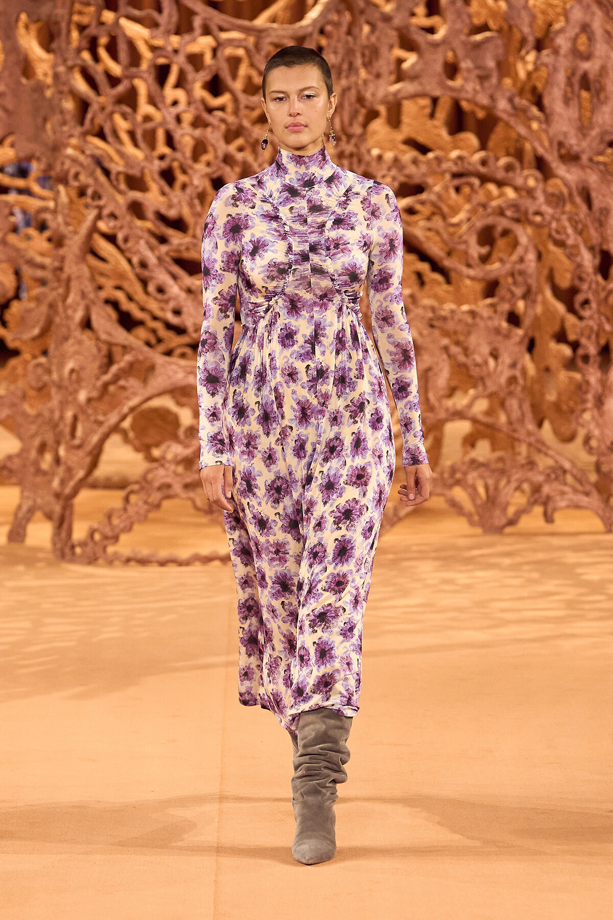 Model walking a runway in a long-sleeve purple floral jumpsuit with a high neck and gathered waist, paired with gray suede boots, orange sculptural backdrop behind.