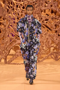 Model walks a fashion runway in a loose, blue‑purple floral jumpsuit with a front zipper, against an orange abstract sculpture backdrop.