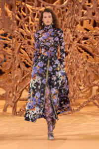Female model wearing a long, high-neck floral dress (blue and purple flowers) walking on a runway with a sculptural brown backdrop.