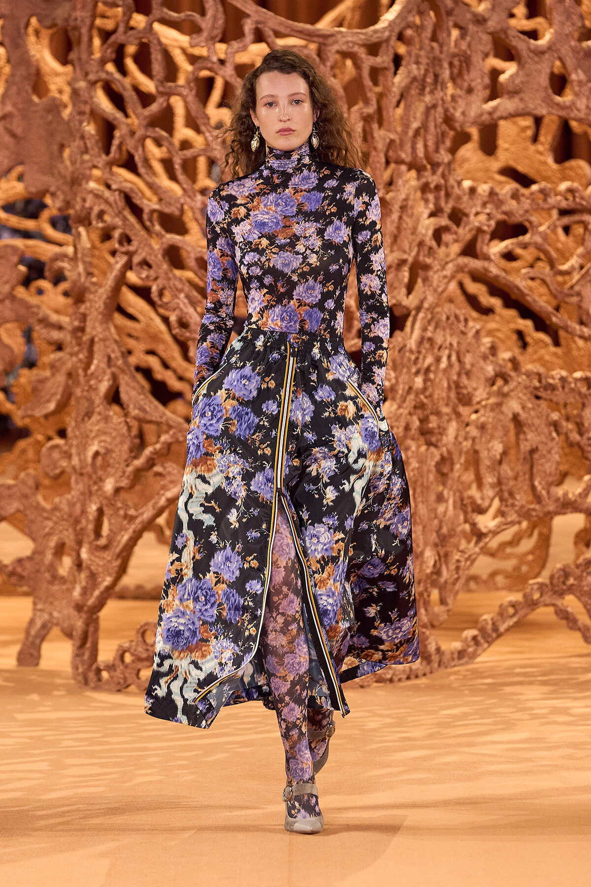 Female model wearing a long, high-neck floral dress (blue and purple flowers) walking on a runway with a sculptural brown backdrop.