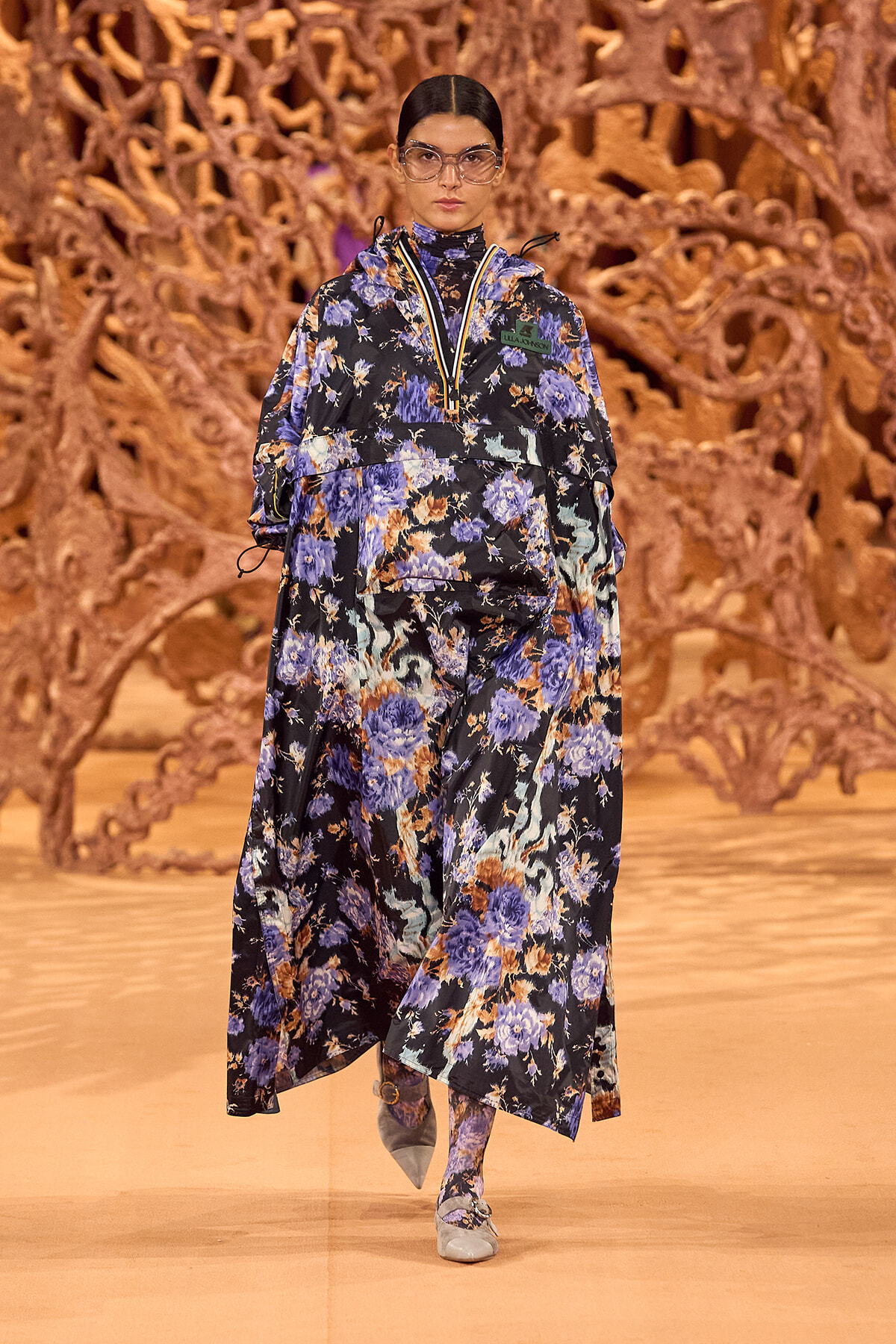 Model walking the runway in a blue‑purple floral print oversized dress with a high collar and matching scarf, wearing clear protective eyewear against an orange sculpted backdrop.