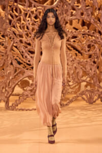 Model walks a runway in a peach knit top and flowing pink skirt, with a tangled brown sculptural backdrop behind her at a fashion show moment.