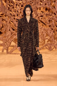 Model walking runway in a dark patterned coat with a belt, carrying a black handbag against an orange sculptural backdrop.