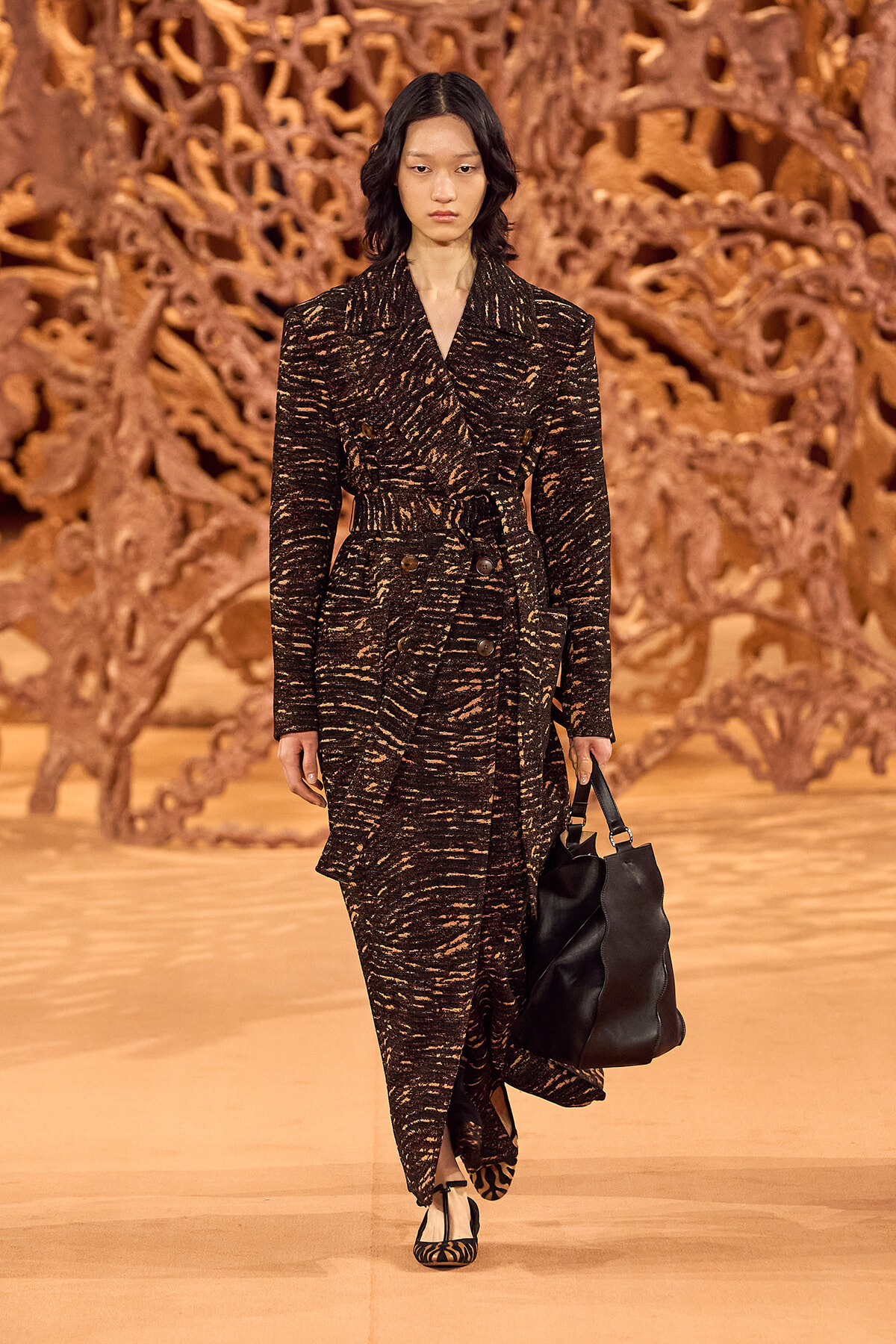Model walking runway in a dark patterned coat with a belt, carrying a black handbag against an orange sculptural backdrop.