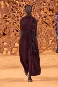 Runway model in a burgundy sleeveless dress with a ruffled side detail walking the catwalk.