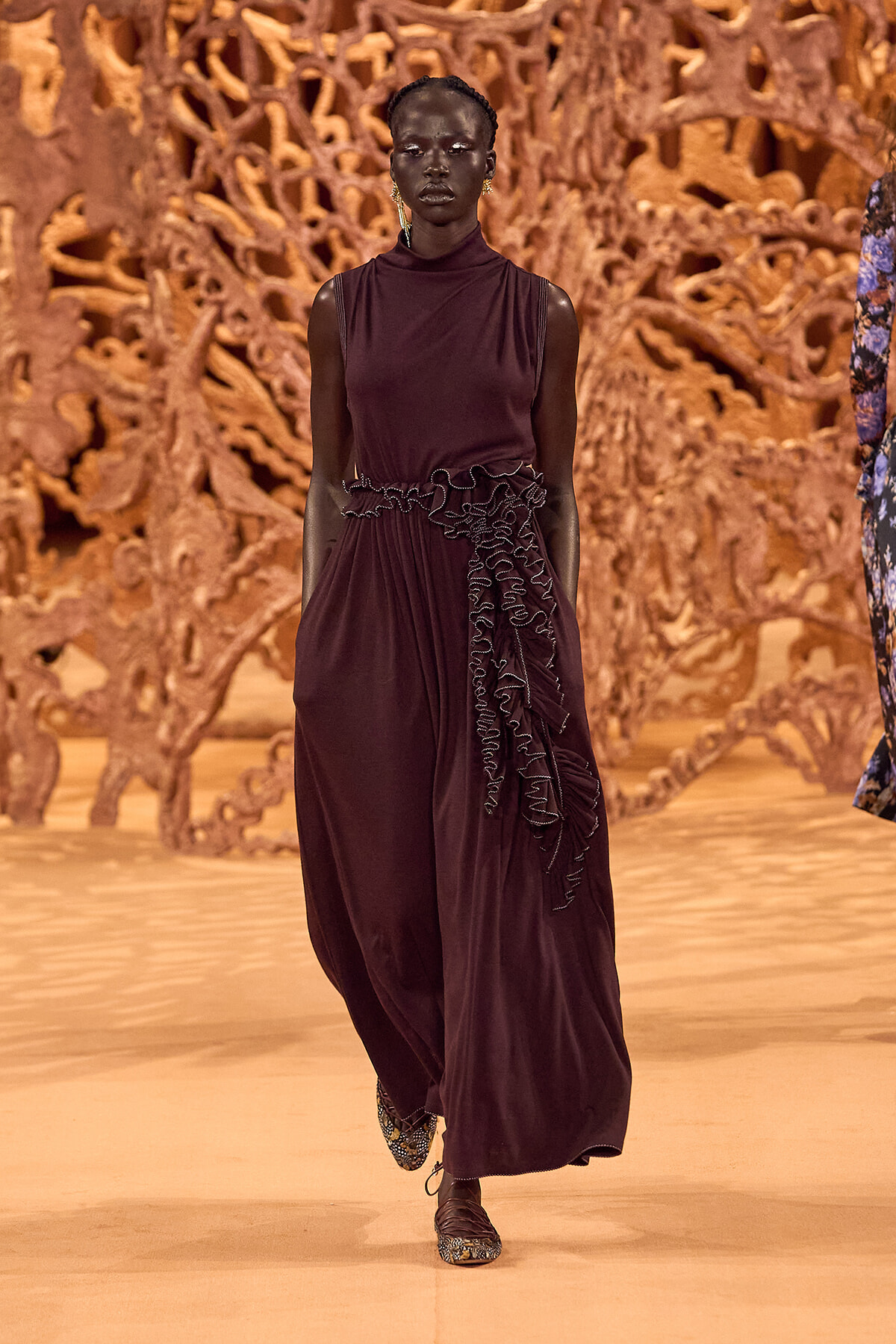 Runway model in a burgundy sleeveless dress with a ruffled side detail walking the catwalk.