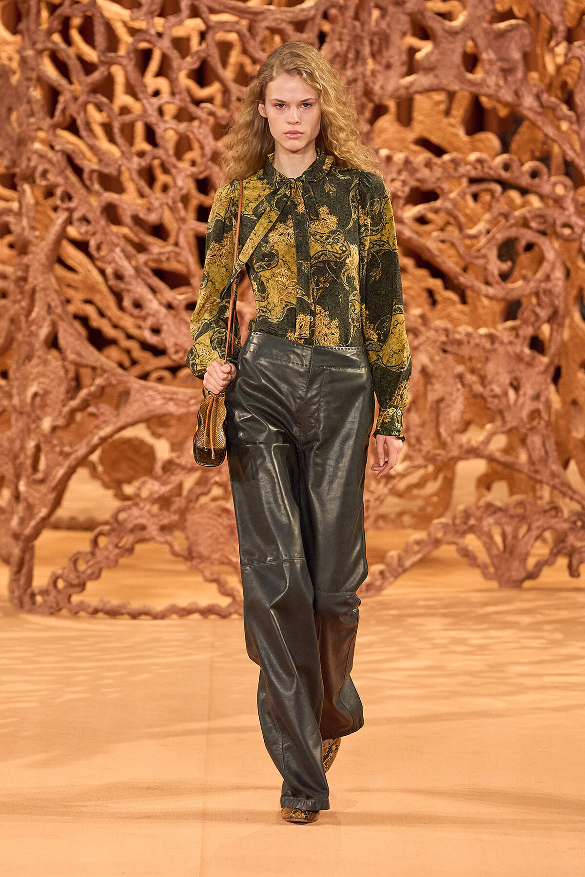 Female model walks a runway in a green paisley blouse and glossy black leather pants against a sculptural brown backdrop, holding a small brown bag.