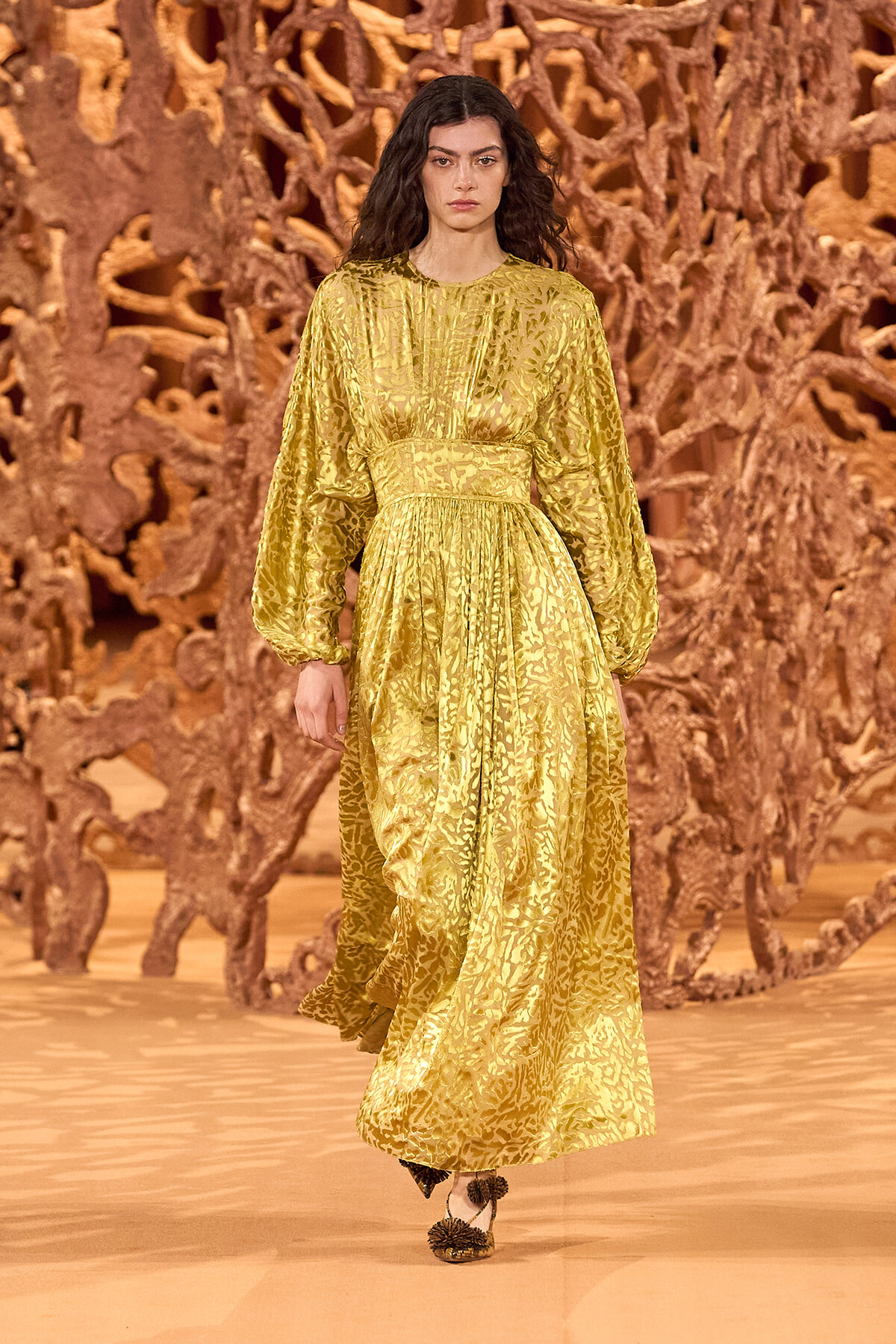 Female model walking the runway in a gold, patterned longue dress with a cinched waist and billowing sleeves, against a brown abstract sculpture backdrop.
