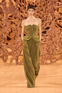 Model walks the runway in an olive pinstriped strapless corset top with matching wide-leg pants, against a sculptural brown backdrop.