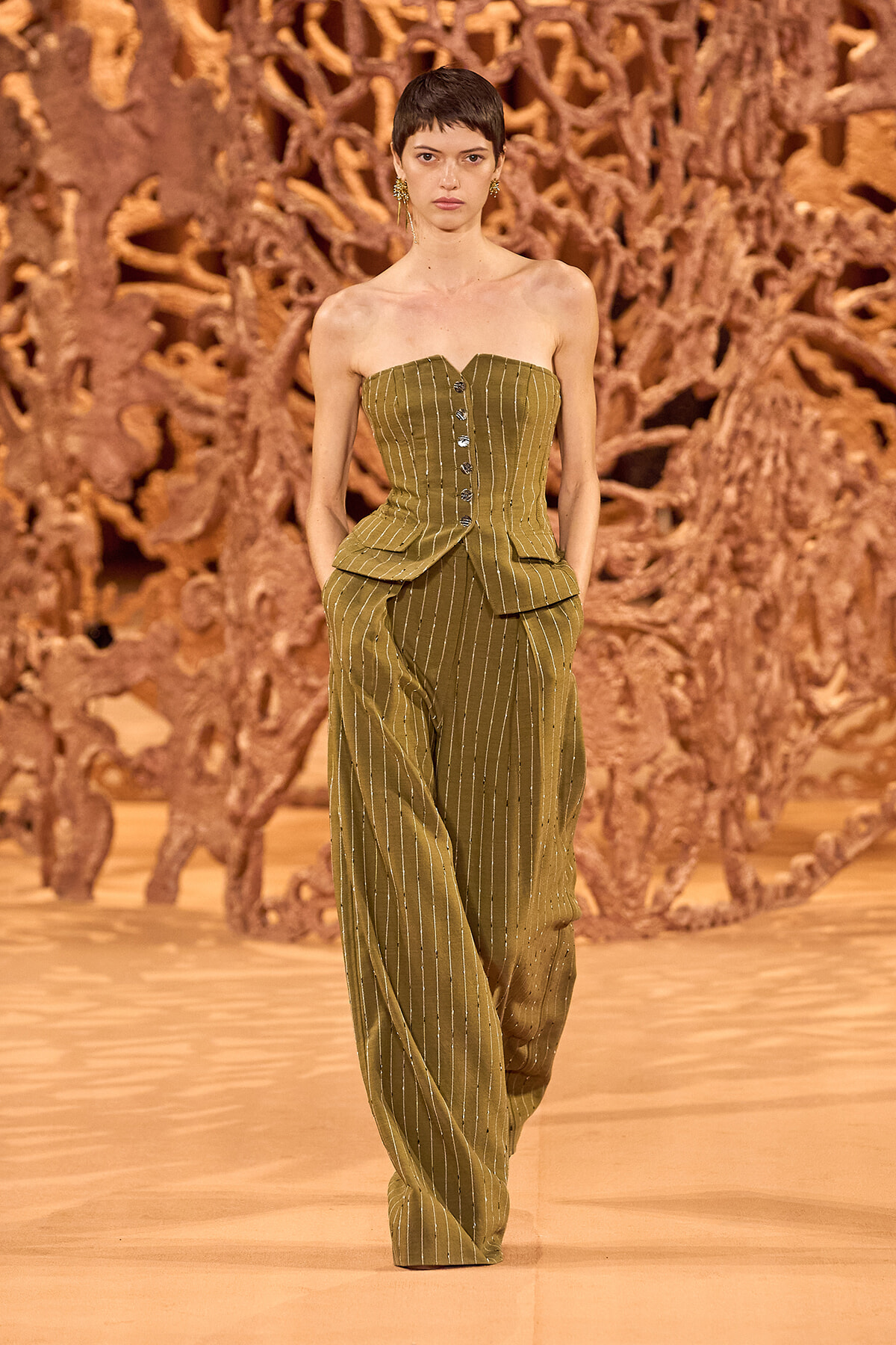 Model walks the runway in an olive pinstriped strapless corset top with matching wide-leg pants, against a sculptural brown backdrop.