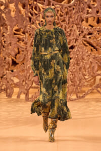 Model walks the runway in a green and gold paisley-print dress with a matching headscarf and snakeskin boots, against an orange sculptural backdrop.