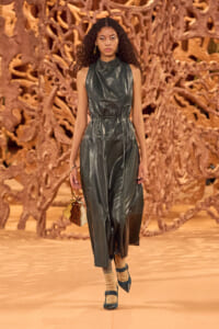 Model walks a runway in a dark green leather sleeveless jumpsuit with a tied waist, against an abstract brown sculptural backdrop.