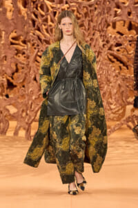 Model walking a runway in a dark green paisley-patterned coat over a black leather dress, with a thin brown crossbody strap visible.