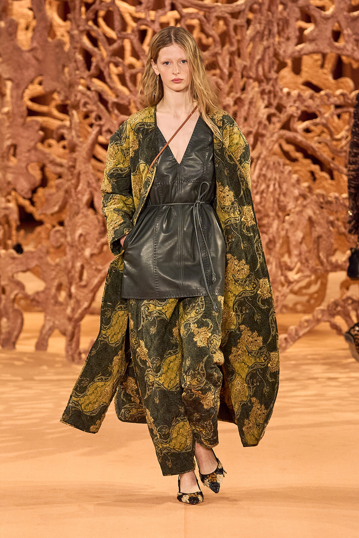 Model walking a runway in a dark green paisley-patterned coat over a black leather dress, with a thin brown crossbody strap visible.