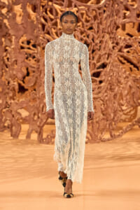 Model walks a runway in a full-length white lace dress with a high neck, intricate floral lattice pattern, against a brown sculptural backdrop.