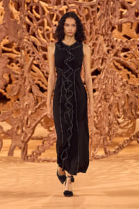 Model wearing a long black sleeveless dress with white piping and a front ruffle, walking the runway against an intricate wooden sculpture backdrop.