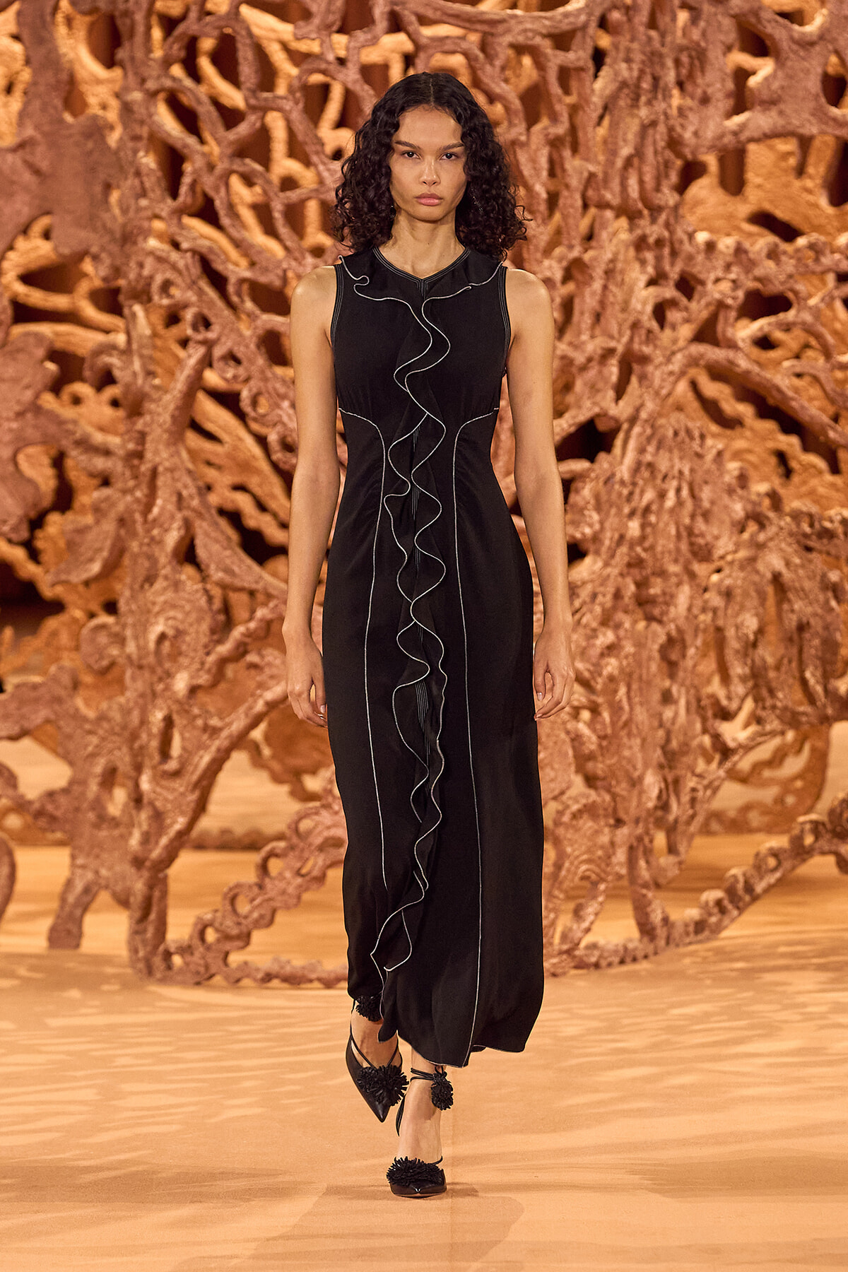 Model wearing a long black sleeveless dress with white piping and a front ruffle, walking the runway against an intricate wooden sculpture backdrop.