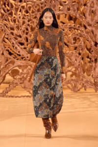 Model walking a fashion runway in an earth-toned paisley dress, tan shoulder bag, and snakeskin boots with a carved wooden backdrop behind.