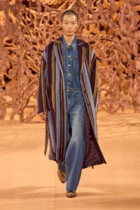 Model walking a fashion show runway in a long multicolored striped coat over a denim jumpsuit, with a gold-toned sculptural backdrop behind.