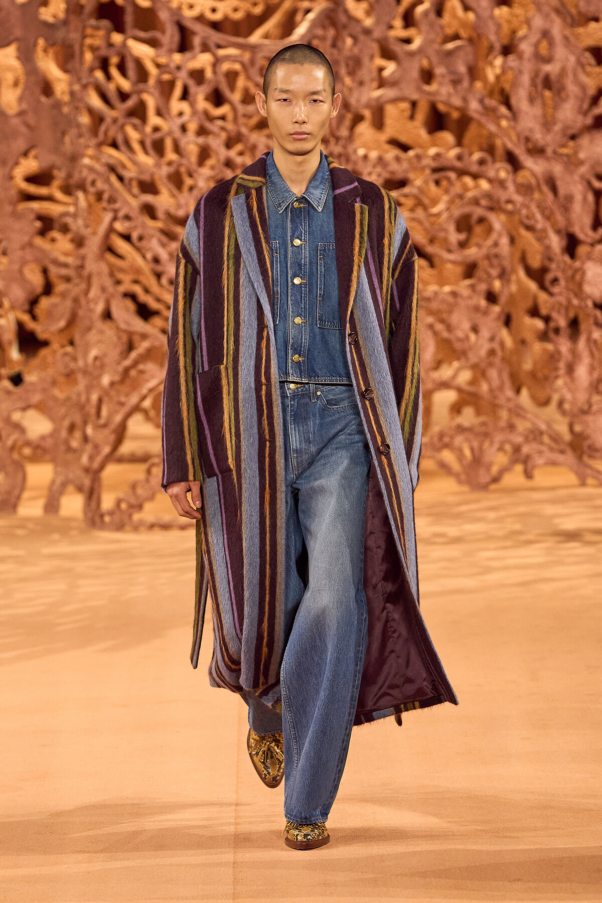 Model walking a fashion show runway in a long multicolored striped coat over a denim jumpsuit, with a gold-toned sculptural backdrop behind.