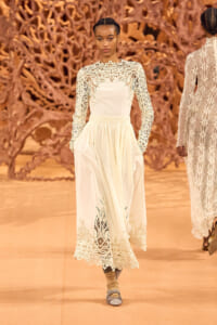 Model walking a runway in a cream lace long-sleeve gown with intricate floral patterns and scalloped hem.