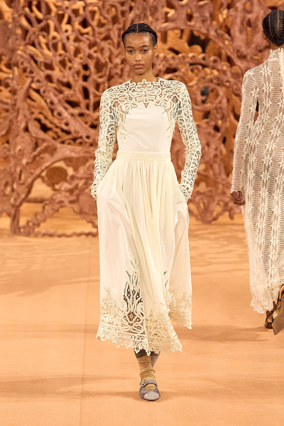 Model walking a runway in a cream lace long-sleeve gown with intricate floral patterns and scalloped hem.